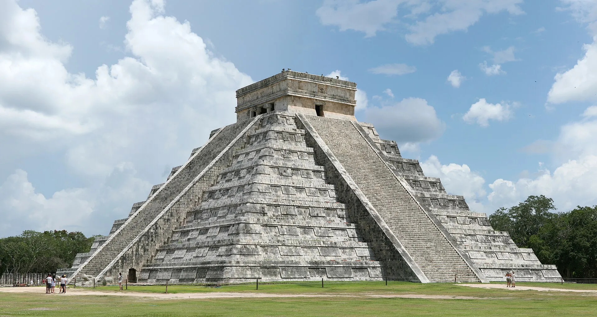 The El Castillo pyramid at Chichén Itzá in Mexico, a giant stone step pyramid with stairs up its sides, on green grass under a blue sky.