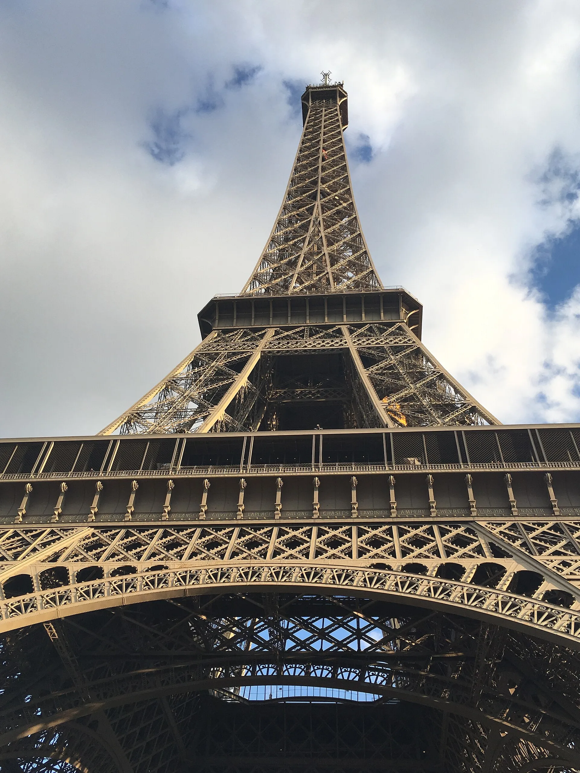 Looking up at the Eiffel Tower's iron lattice into a cloudy sky