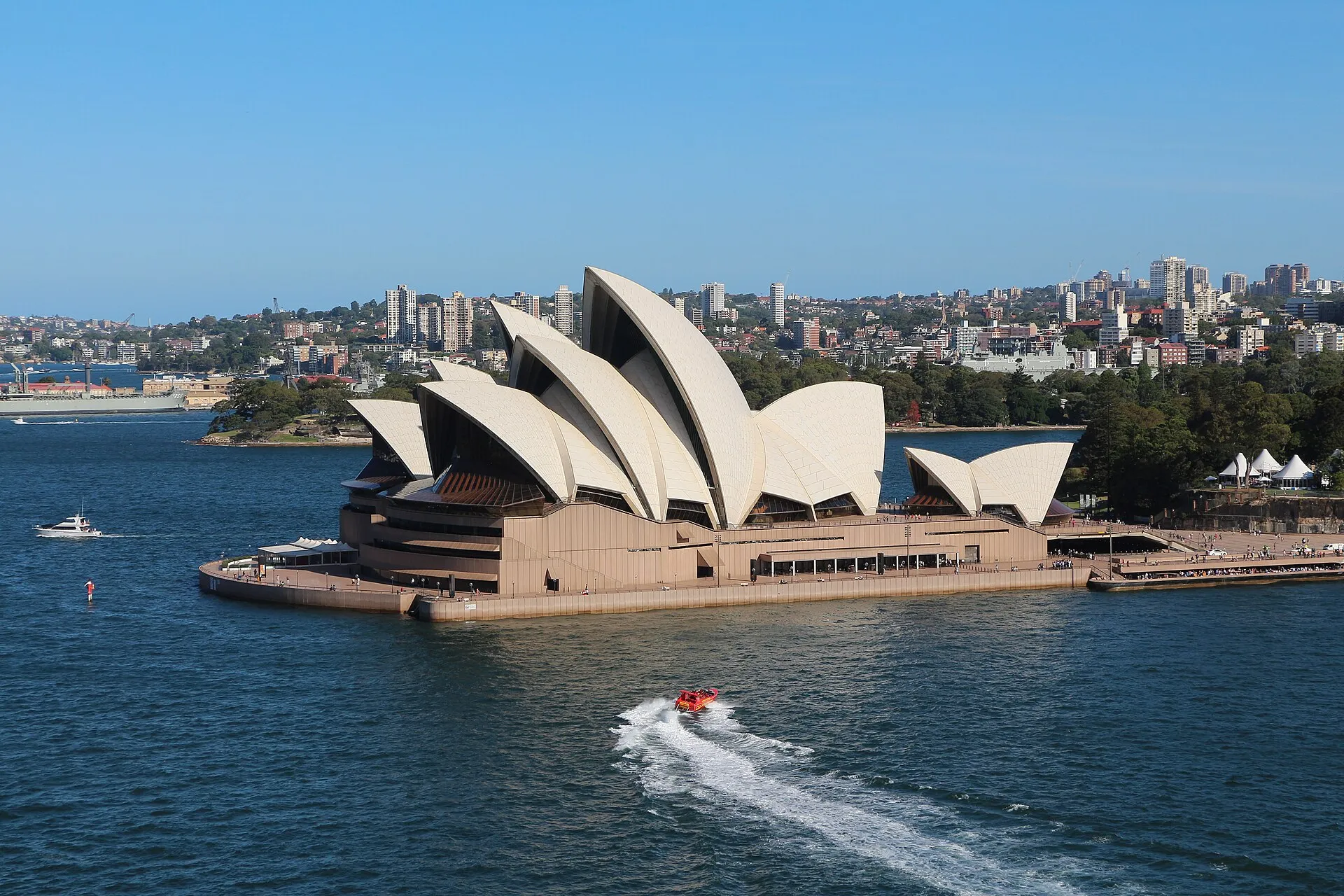 The Sydney Opera House beside the blue harbour on a sunny day, with its white roof shaped like big sails.